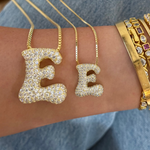 Iced jumbo and medium jumbo letter E necklaces held over a wrist with gold bangles