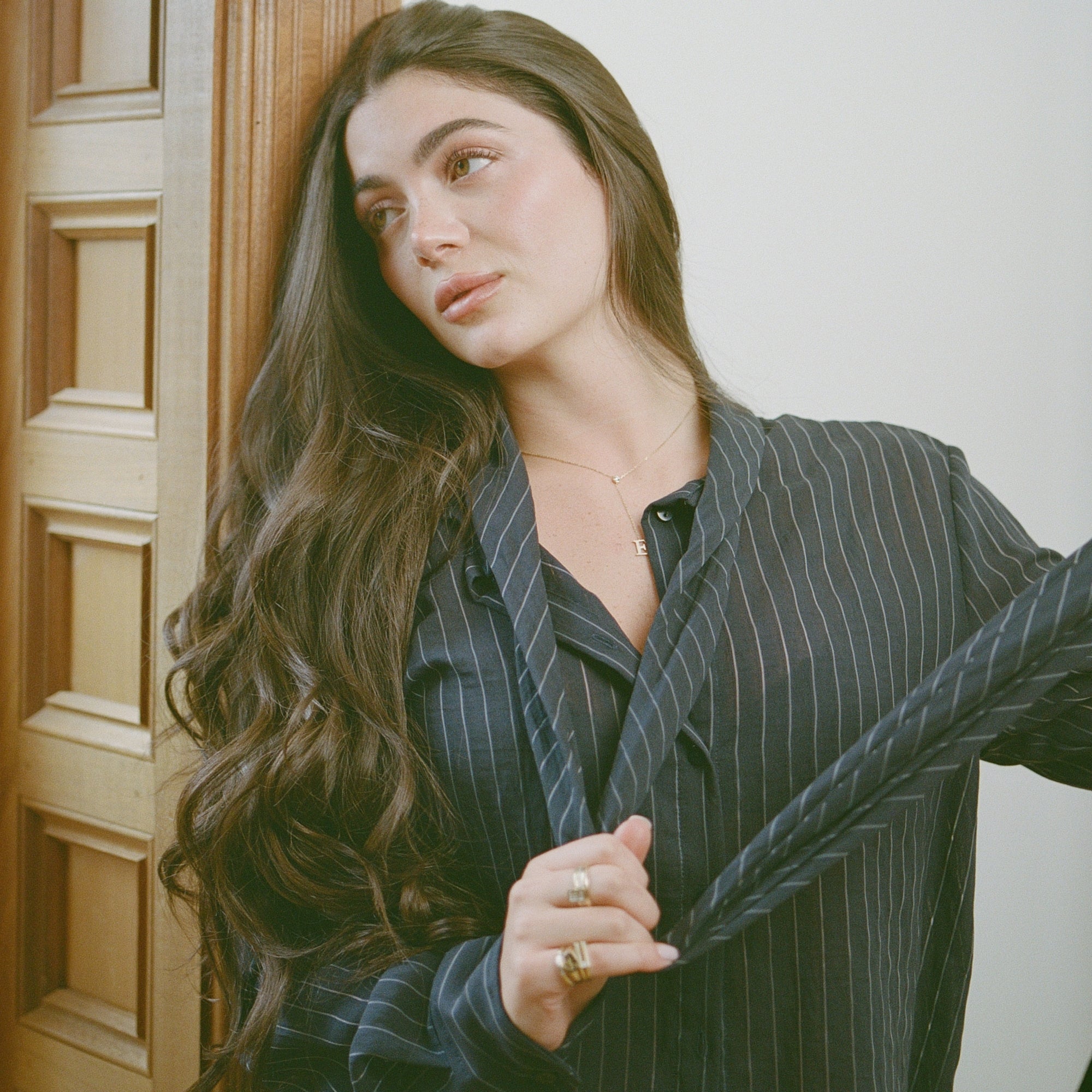 Model in a pinstripe shirt wearing the Timeless Lariat Letter Necklace by a wooden door